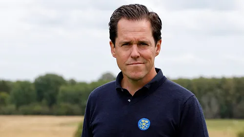 Man i marinblå poloskjorta med SGF:s logotyp, utomhus med golfbana i bakgrunden.