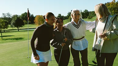 Fyra kvinnor i olika åldrar ler och håller om varandra på en golfbanaa.