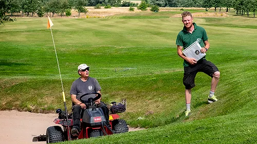 Banpersonal på traktor och man som står vid en bunkar på golfbana med flagga i bakgrunden