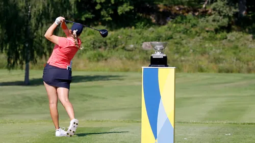 Golfspelare slår ett utslag från tee. I förgrunden syns en blågul piedestal med en pokal.