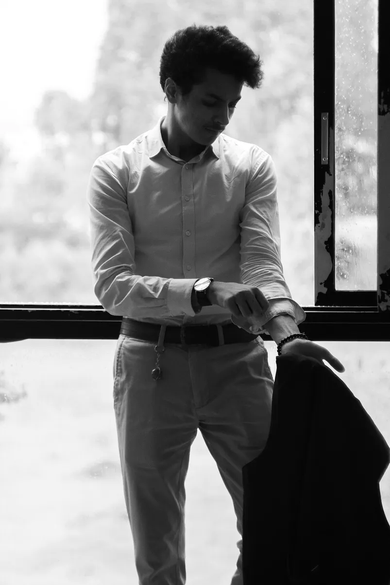 Black and white photo of a man standing by a window while rolling up his shirt sleeve