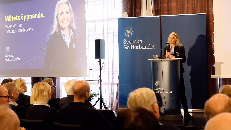 En kvinna på en scen vid ett podium, inför publik som sitter och lyssnar. 