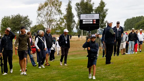 Ung golfspelare håller upp resultattavla på golfbana med åskådare och officiella personer i bakgrunden.