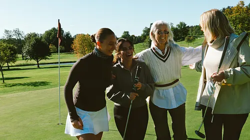 Fyra kvinnor i olika åldrar ler och håller om varandra på en golfbanaa.