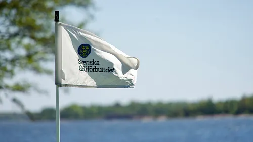 Svenska Golfförbundets flagga med vit bakgrund och blå emblem, vajar i vinden vid en sjö
