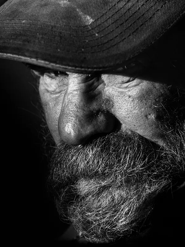 Black and white close-up of an elderly bearded man wearing a cap, lit dramatically from the side