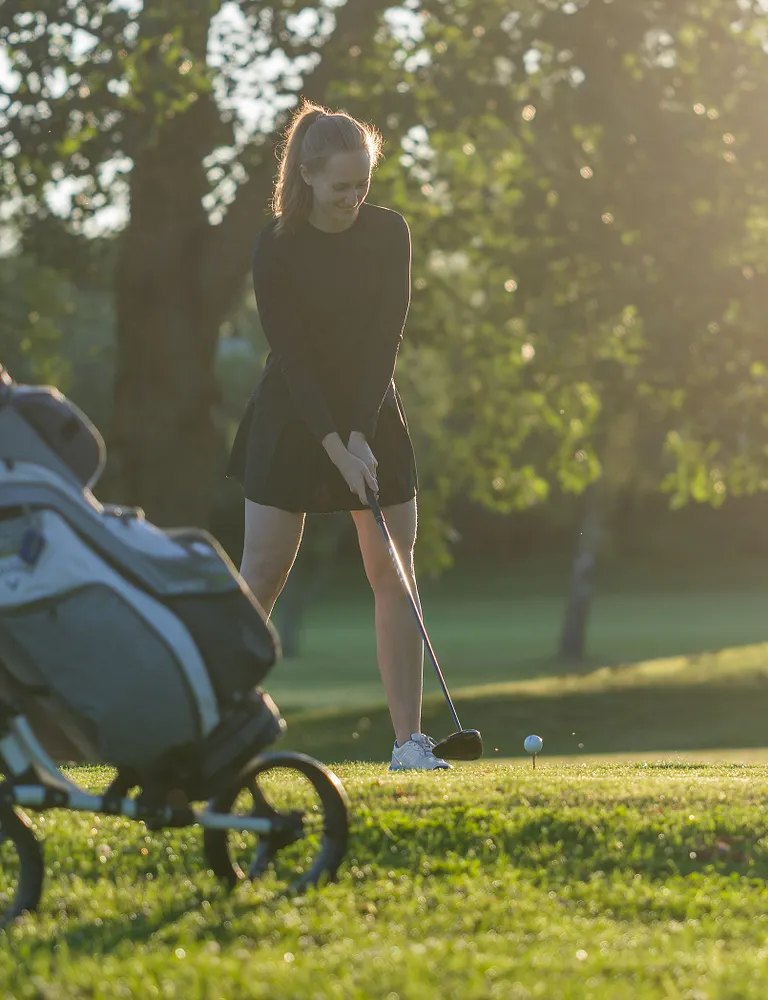 Kvinna står med sin driver på tee på en golfbana, i solljus med träd i bakgrunden.