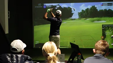 En tonåring slår ett slag i en golfsimulator inomhus. Tre kompisar tittar på.