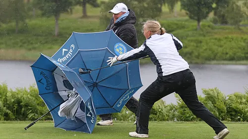 Två golfspelare kämpar med ett blått paraply i kraftig vind på golfbanan.