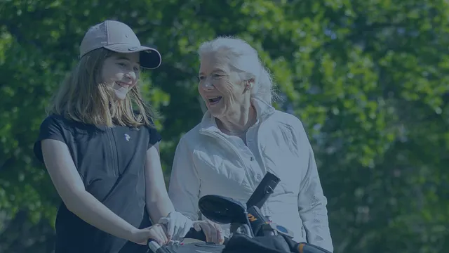En flicka i 12-årsåldern med sin farmor, ler och går på golfbanan med en golfvagn.