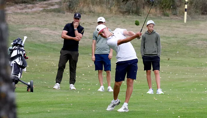 Juniorspelare slår ett slag på en golfbana medan kompisarna observerar.